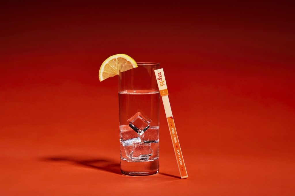 a photo of a clear glass filled with clear liquid and ice and a citrus wedge on the edge against a red backdrop the myhi stir stick is leaning against the glass