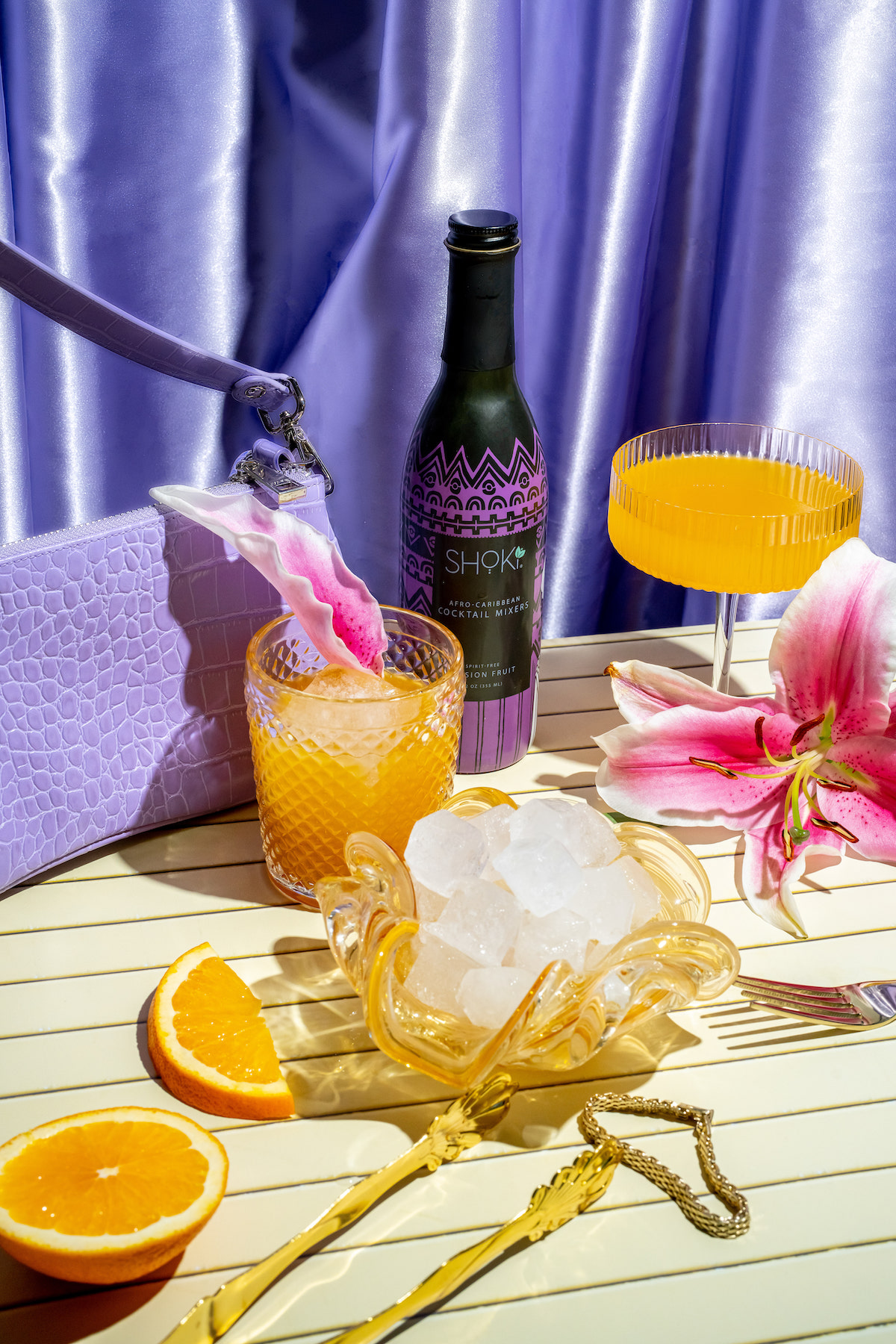Shoki display of orange beverages in cocktail glasses adorned by pink lilies against a lavender satin curtain