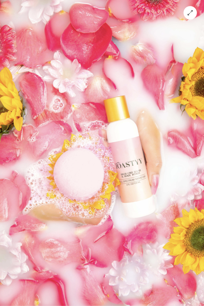 aeerial photo of a white lotion bottle with a light pink label and a round light pink bath bomb surrounded by bright pink and yellow flowers submerged in water over a white backdrop