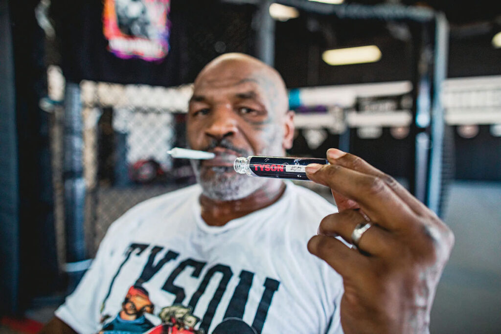 Mike Tyson with a prerolled joint in his mouth holding the preroll tube in the foreground of the image