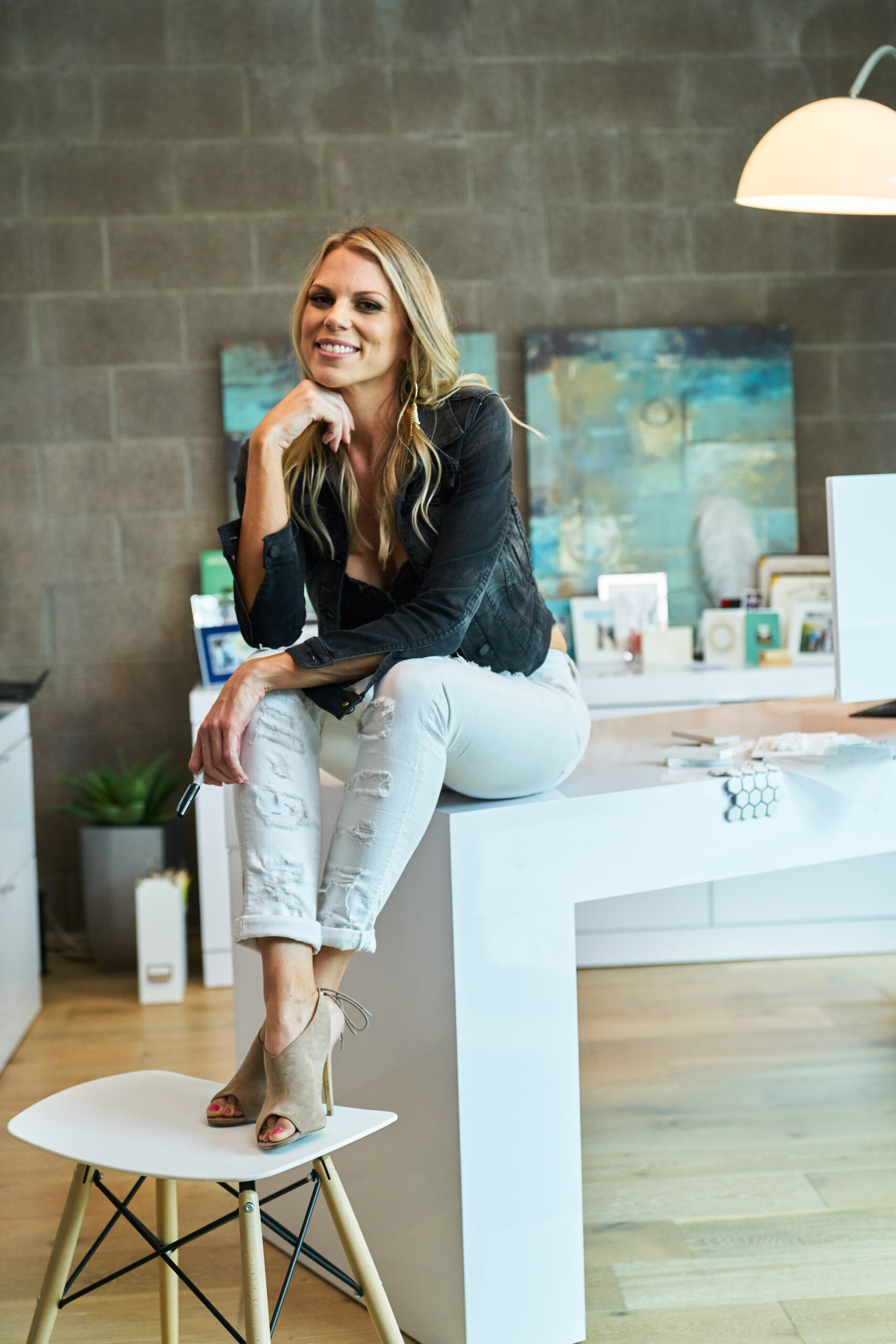 photo of blond woman with long hair leaning on her hand sitting on a table she is smiling and wearing a black shirt white pants and beige heels
